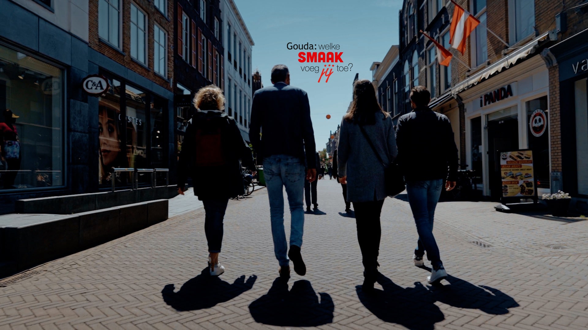 Op de foto staan 4 collega's met hun rug naar het beeld die in de binnenstad van Gouda lopen. In het midden het logo Gouda: welke smaak voeg jij toe?