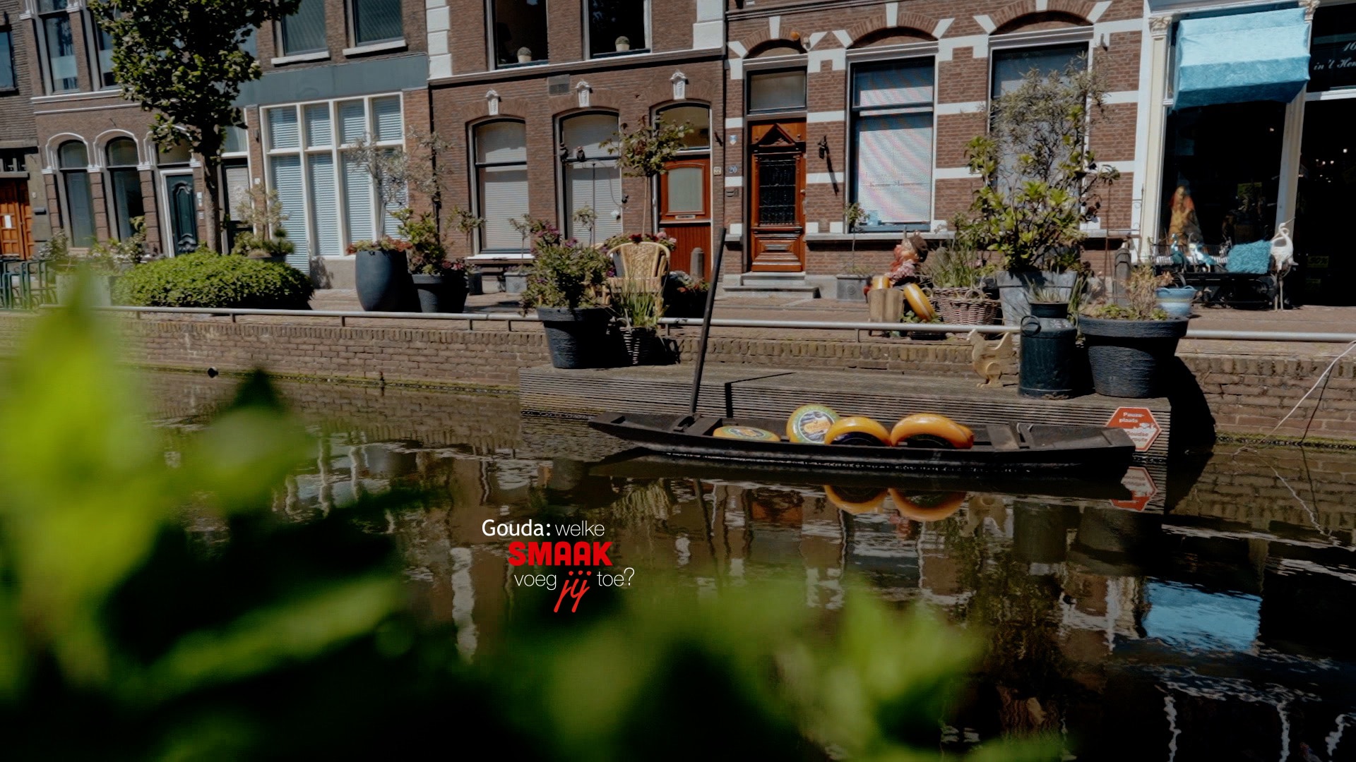 Gracht in Gouda met in het water een bootje met Goudse kazen. In het midden het logo Gouda: welke smaak voeg jij toe?